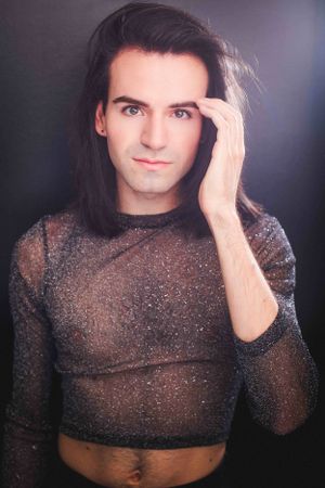Headshot of an actor in a translucent sparkly black shirt tucking their hair behind their ear.