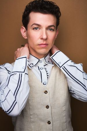 Headshot of an actor in a structured white shirt and beige vest fixing their collar.