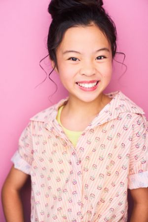Headshot of an adorable young girl in a patterned shirt with hair up in a bun and tendrils falling down against a bright pink background.