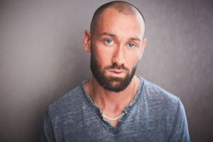 Headshot of a man with a bead in a grey shirt with a chain.