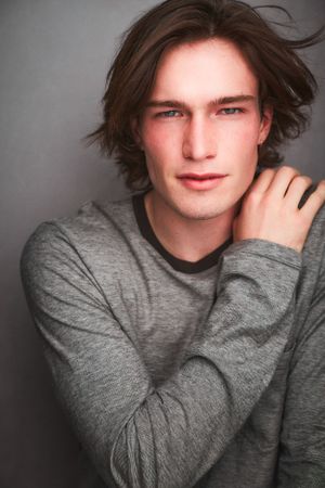 Headshot of a beautiful man with long hair reaching up to his should in a grey shirt.