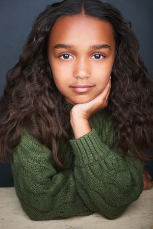 Headshot of a beautiful young girl with long curly black hair wearing a green sweater and resting her head on her hand.