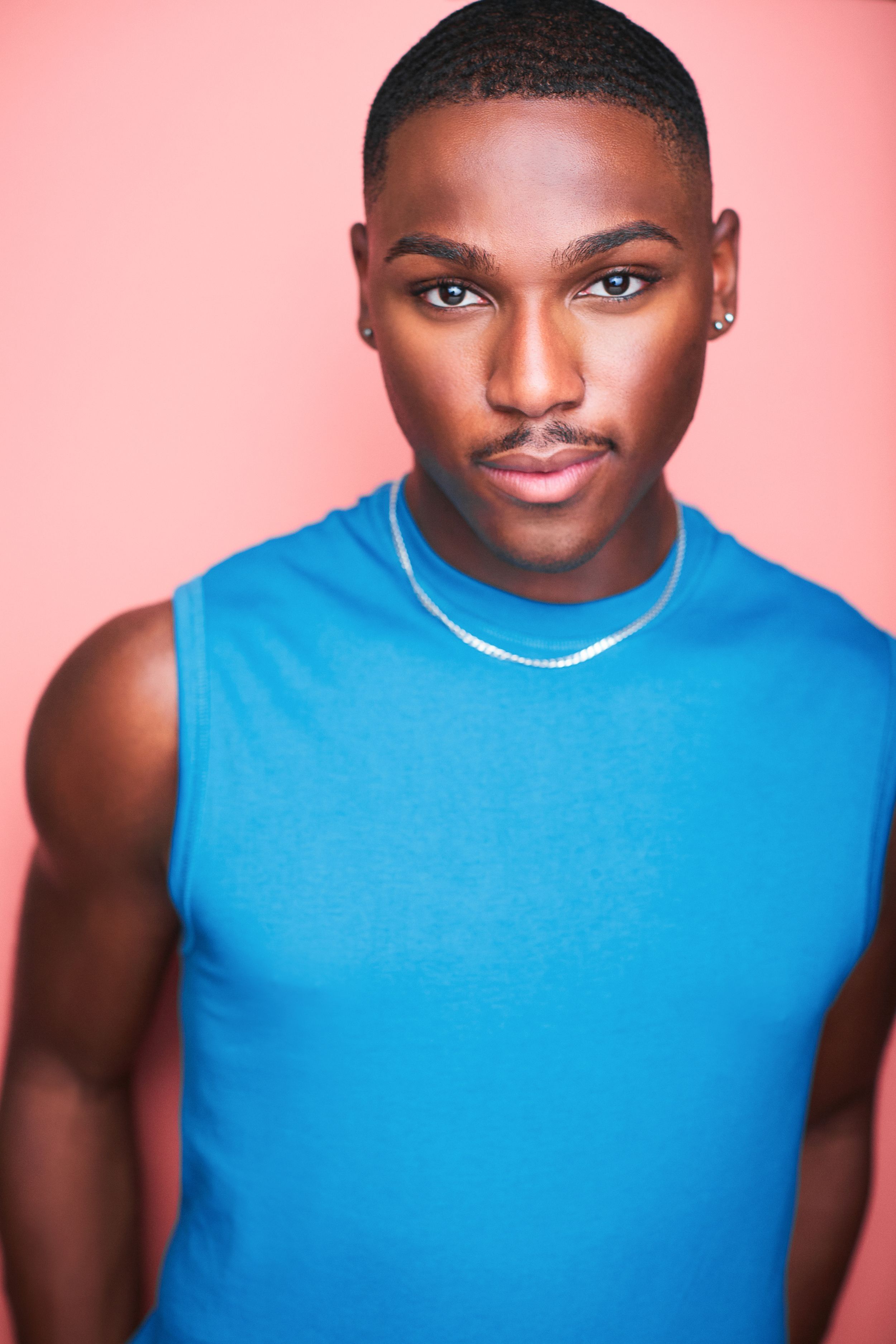 Headshot of a beautiful man in a blue tank top with a chain against a peach background.
