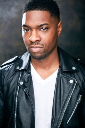 Headshot of a man smirking in a white shirt and leather jacket against a dark background.