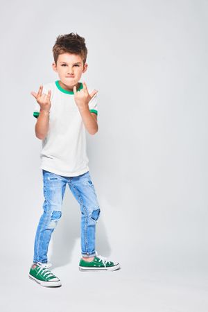 Boy in white T-shirt, ripped jeans, green sneakers, striking a rock-n-roll pose.