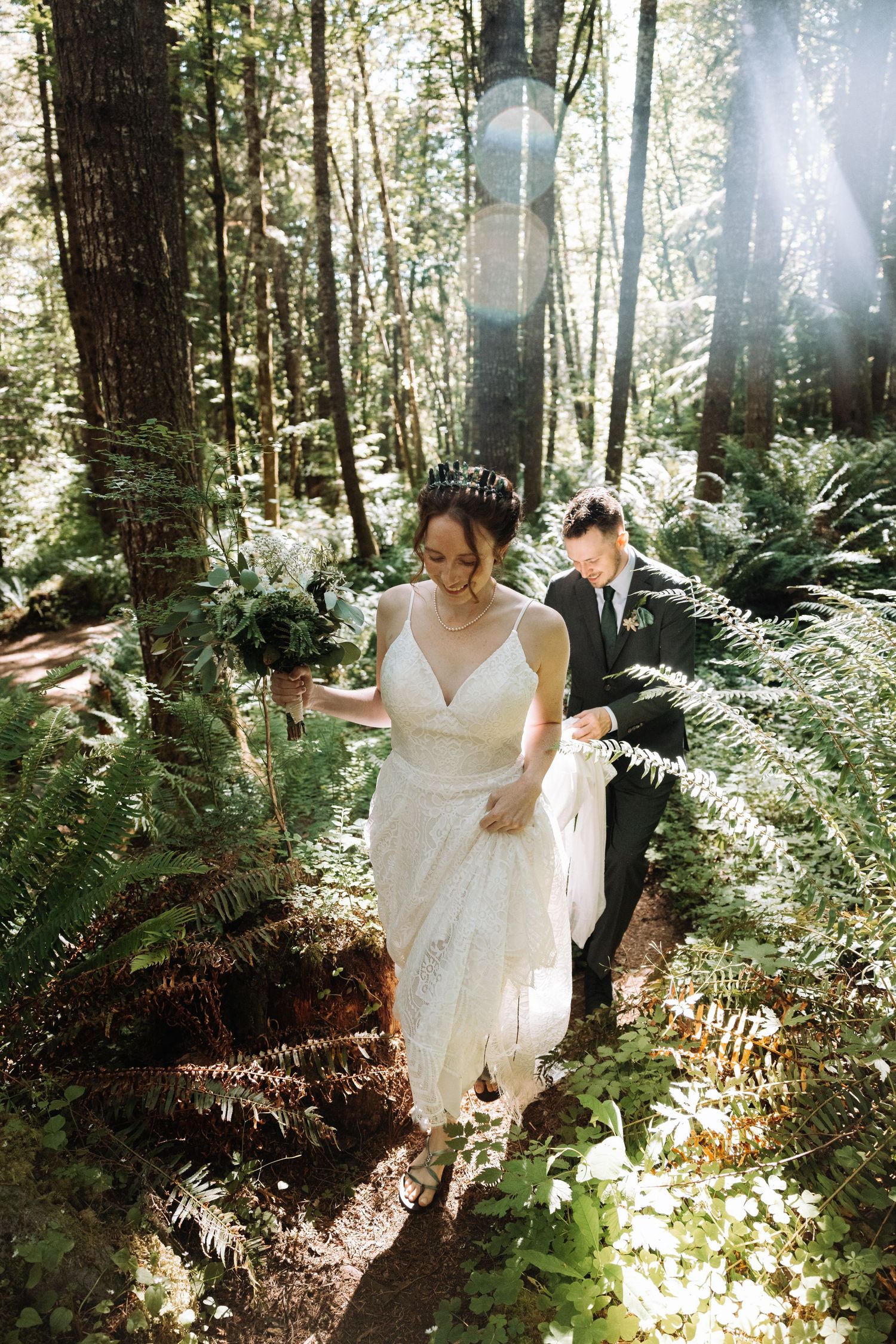 A wedding couple stands together on a forest path surrounded by towering evergreen trees and lush ferns.