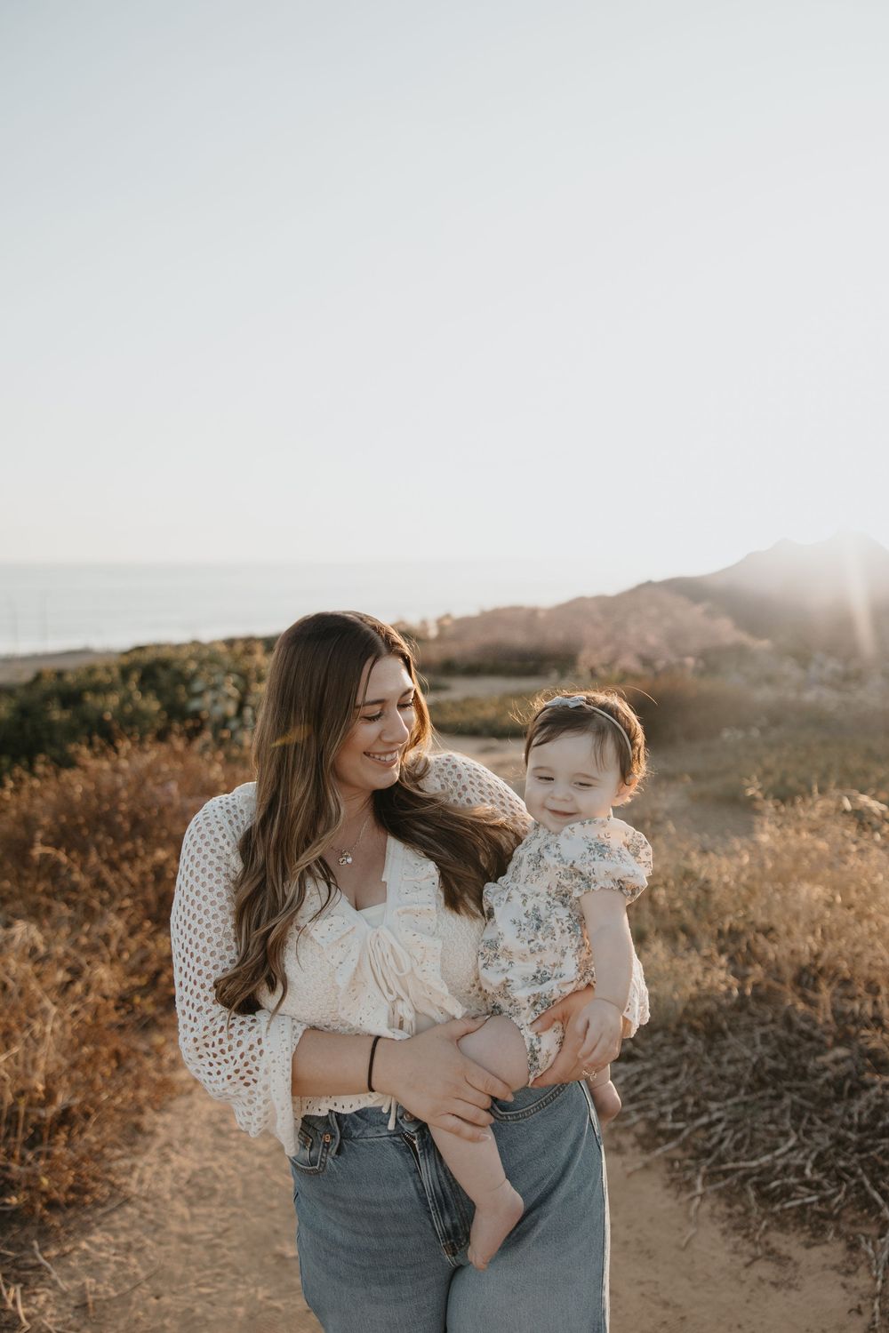 A casual outdoor portrait of a mother and baby at sunset in a scenic mountainside location.