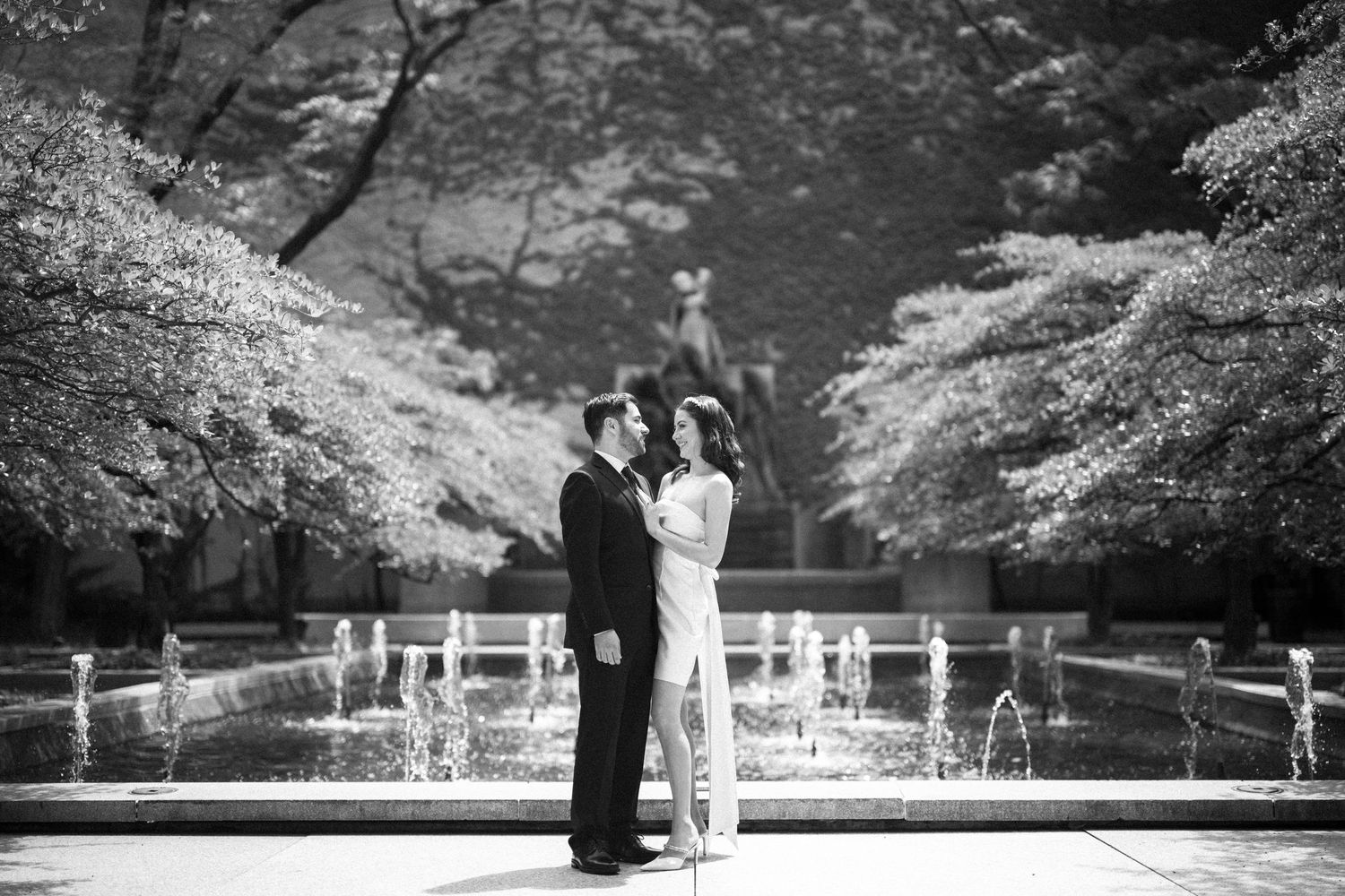 Elegant couple stands together against a backdrop of trees and fountains in black and white.