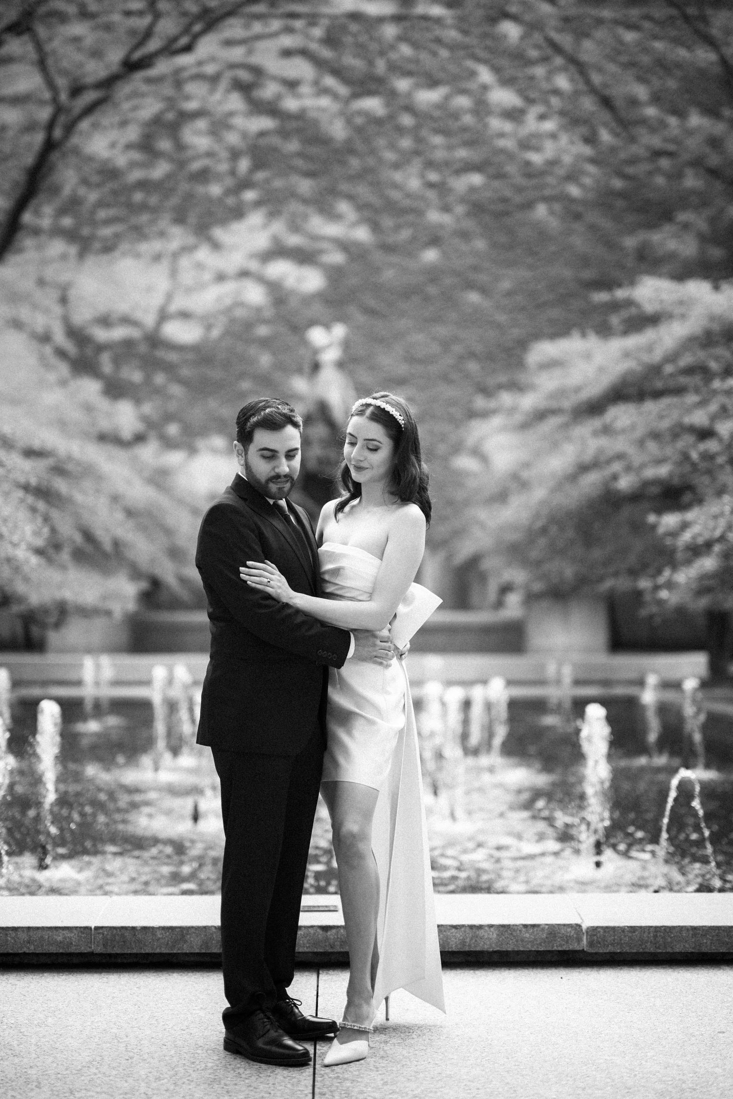 A romantic couple pose in formal attire by a fountain in a garden setting.