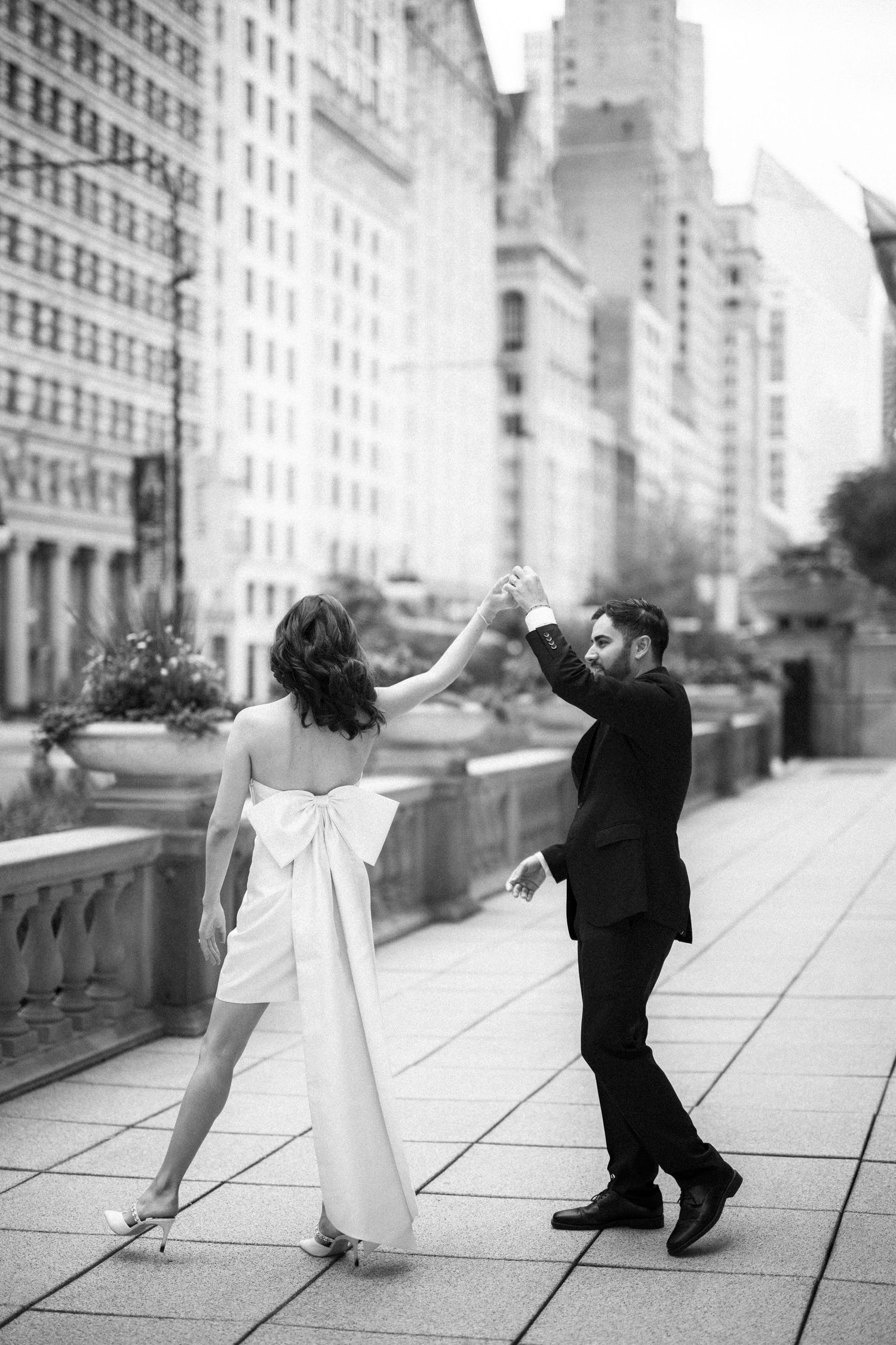 A couple in formal attire dances together on a balcony with city buildings in the background.