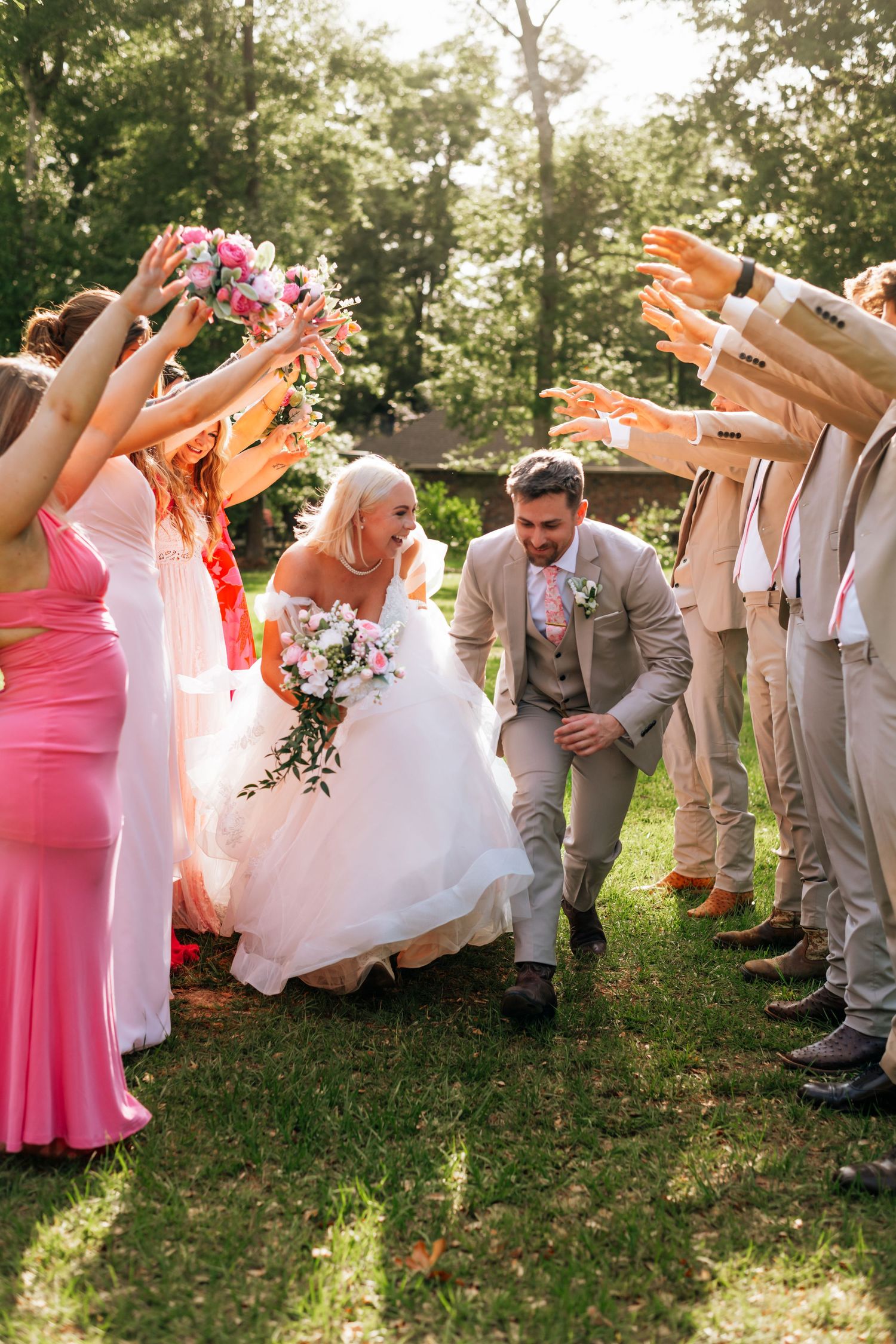 Wedding party celebrates as newlyweds duck under raised arms in outdoor garden setting.