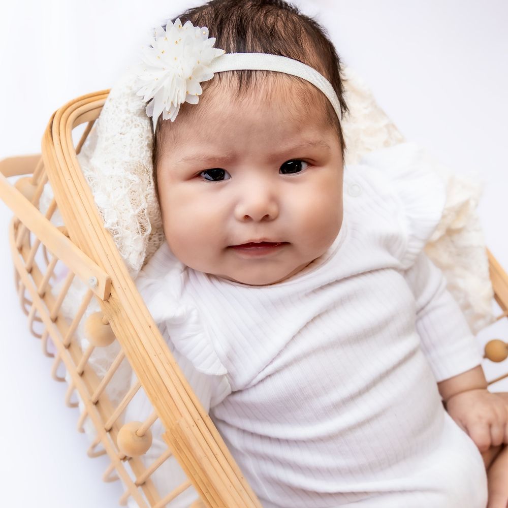 A newborn baby in a white outfit and floral headband rests in a wicker bassinet against a white background in Connell, Wa.