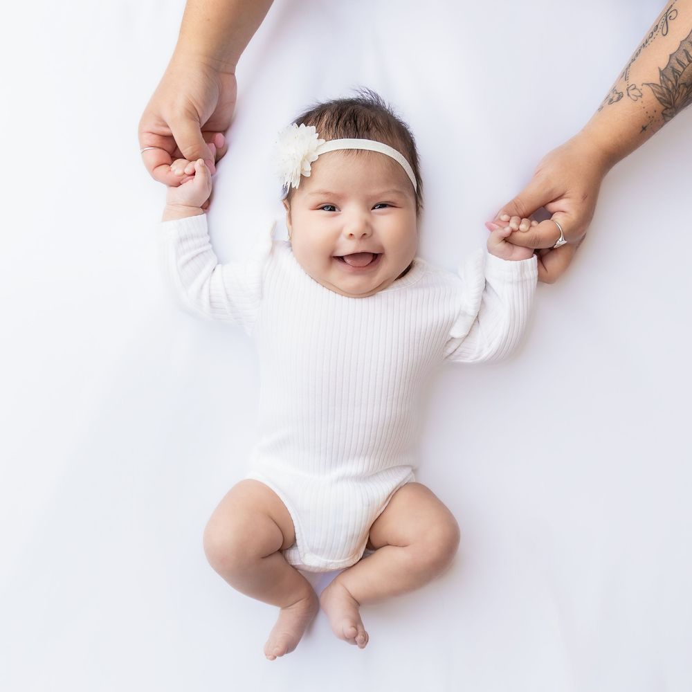 Smiling baby girl in white ribbed onesie and floral headband held by parents on white background in Connell, Wa.
