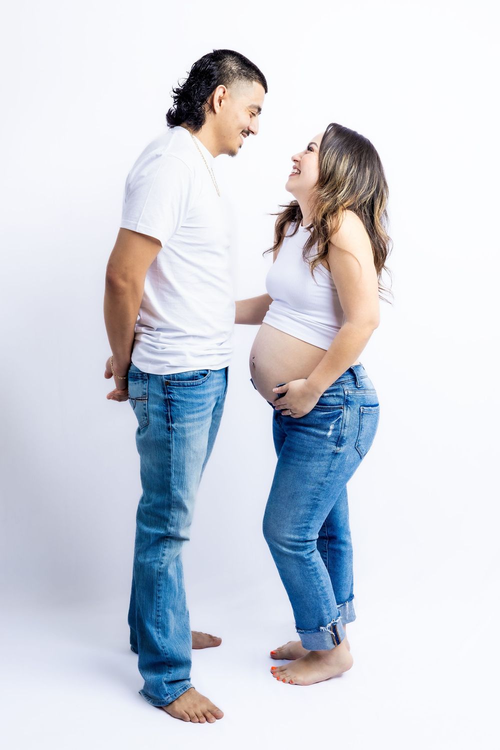 Happy pregnant couple in white tops and jeans smiling at each other, woman cradling baby bump in studio in Connell, Wa.