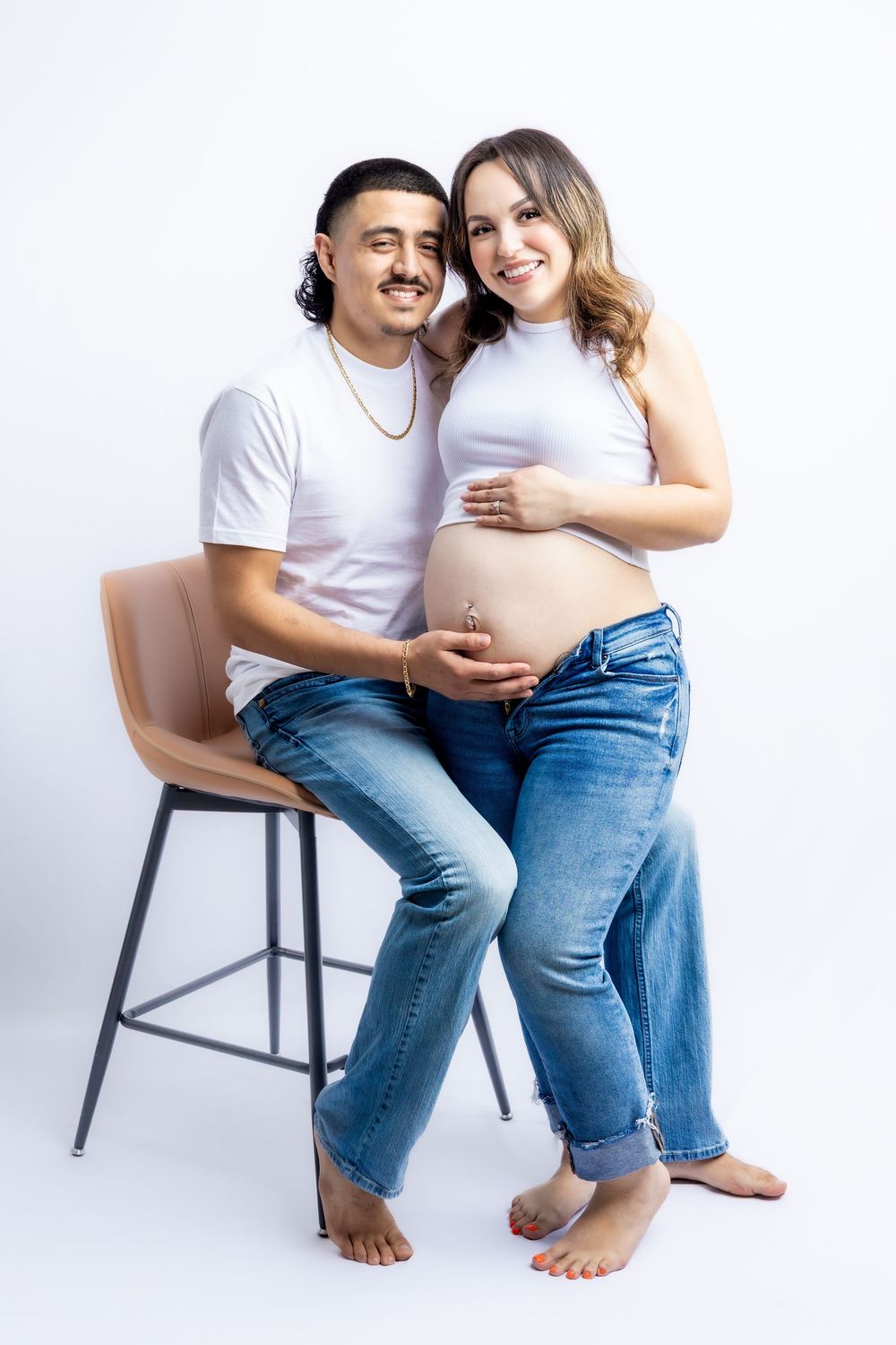 Happy couple in white shirts and jeans posing together, woman showing pregnant belly in studio setting in Connell, Wa.
