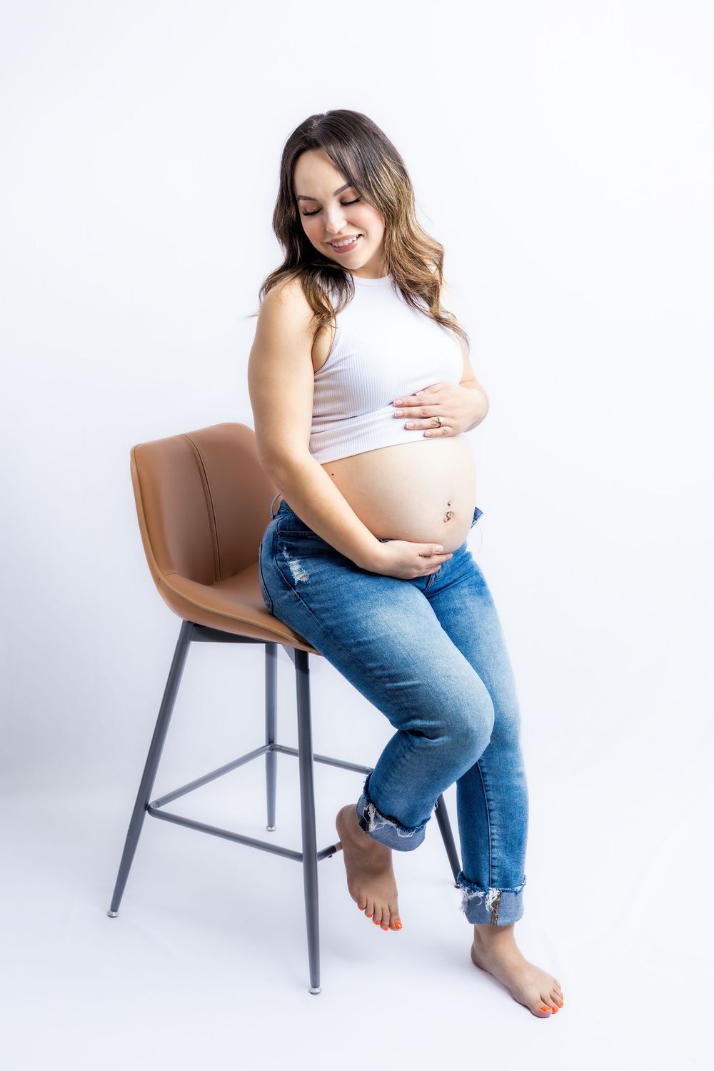 Smiling pregnant woman in white crop top and jeans sitting on a bar stool, cradling her baby bump against white background in Connell, Wa.