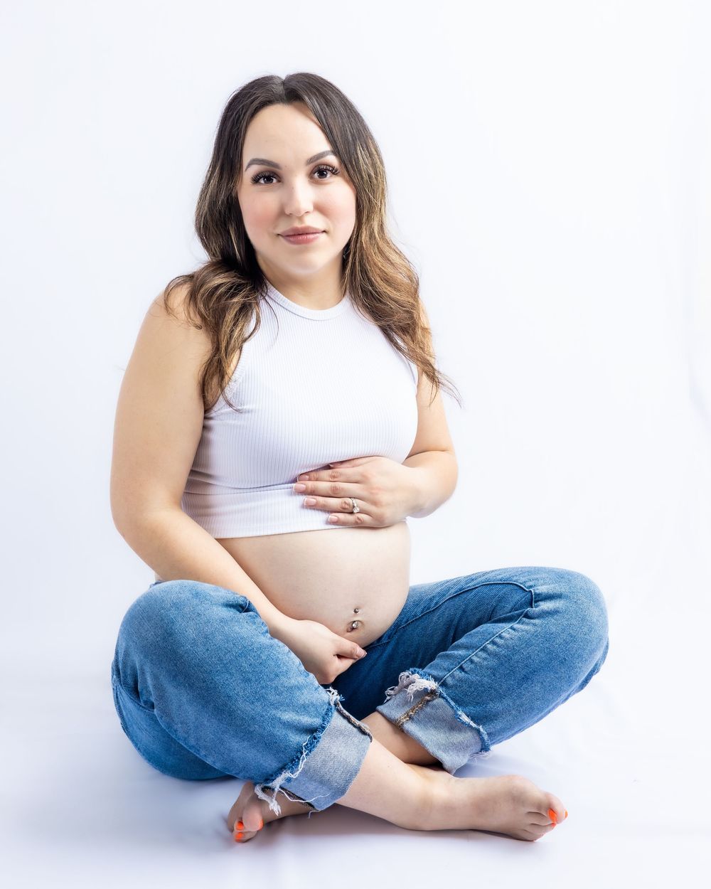 Pregnant woman sitting cross-legged on white background, wearing white crop top and jeans, cradling baby bump in Connell, Wa.