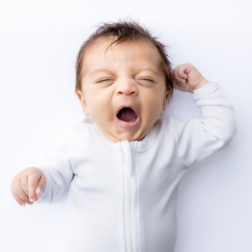 A newborn baby in a white zip-up onesie yawning widely with eyes closed and one arm raised on a white background in Connell, Wa.