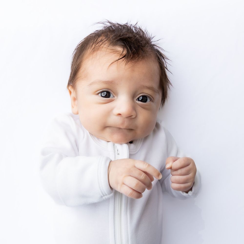 Adorable baby in white onesie lying down, looking up with bright eyes and a sweet smile on white background in Connell, Wa.