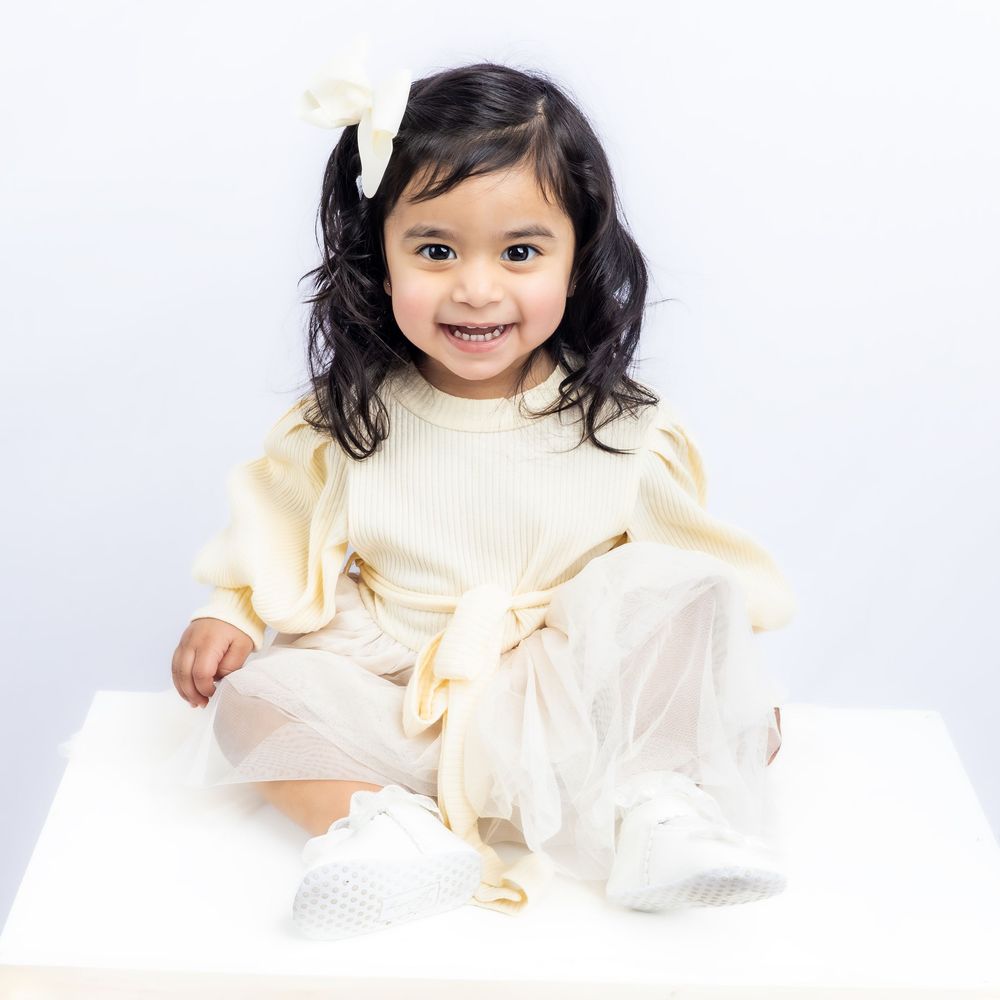 Smiling toddler girl in cream outfit with tulle skirt and white bow, sitting on white surface against light background in Connell, Wa.