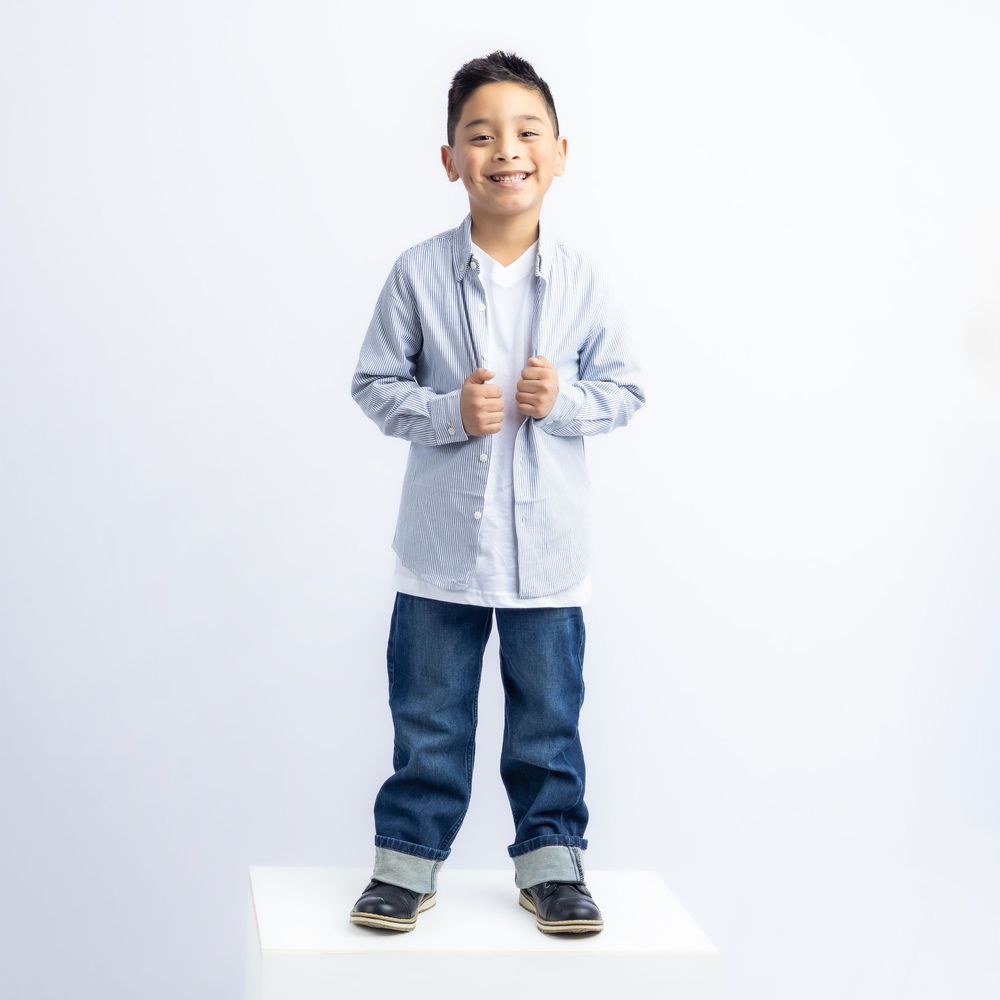 Smiling young boy wearing a blue striped button-up shirt, white tee, dark jeans, and navy shoes on a white platform in Connell, Wa.