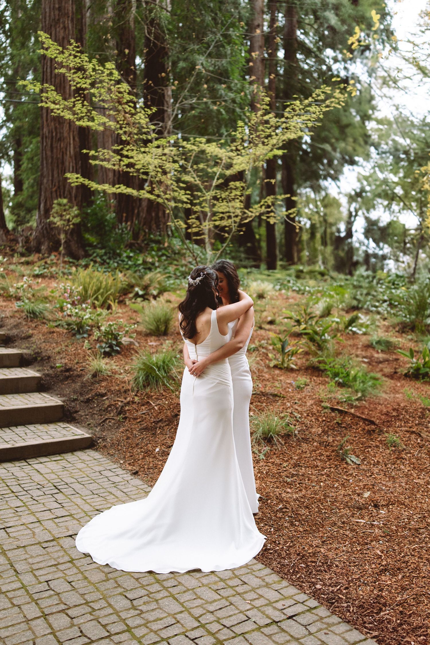 Two people in white dresses embrace on a stone path surrounded by tall redwood trees.