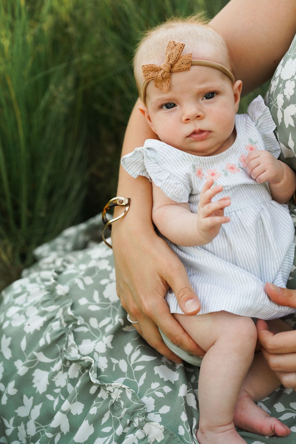 A baby in a white dress sits on a floral patterned cushion while being held by adult hands.