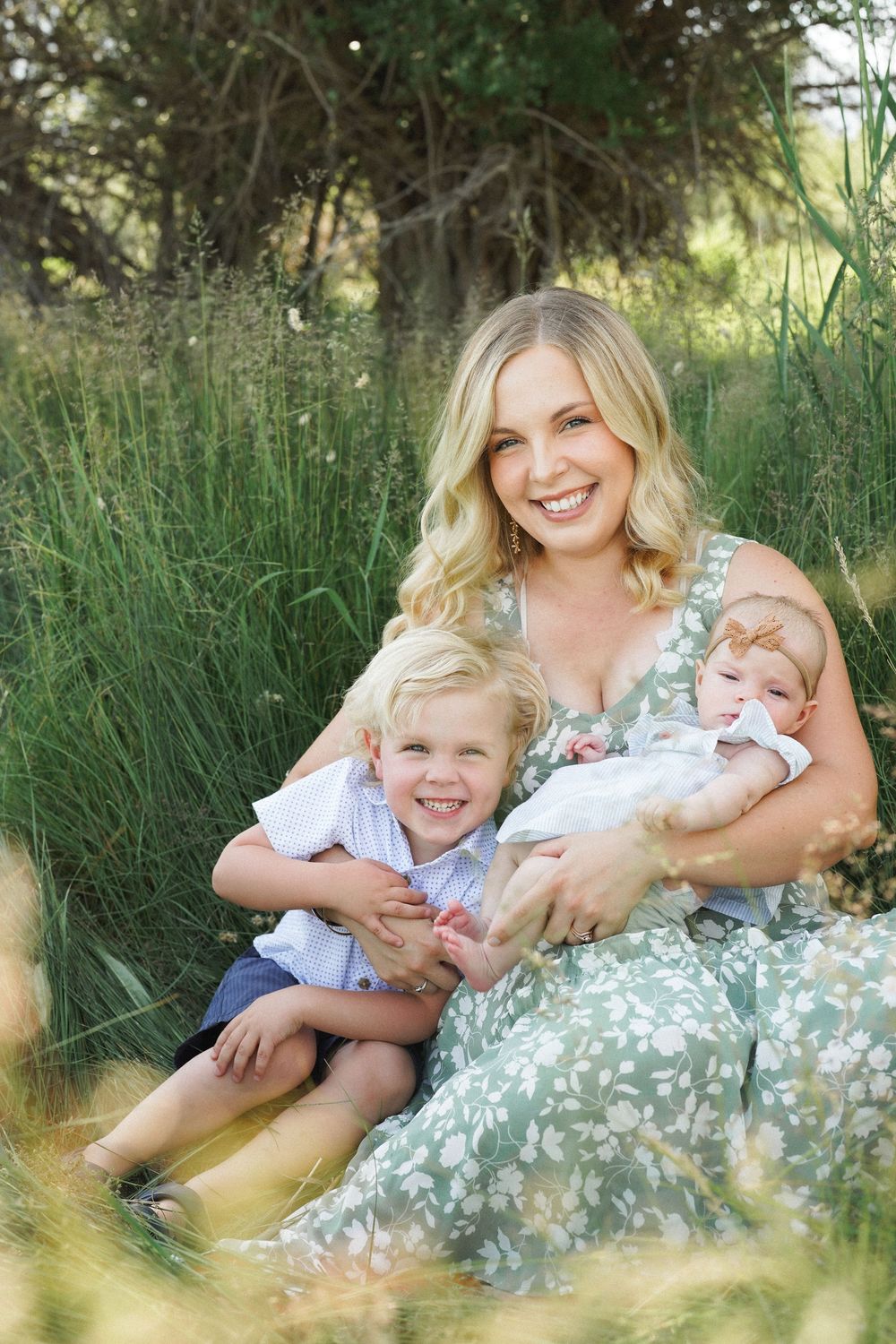A family lying together in tall grass during a sunny outdoor photo session wearing coordinated blue and white outfits.