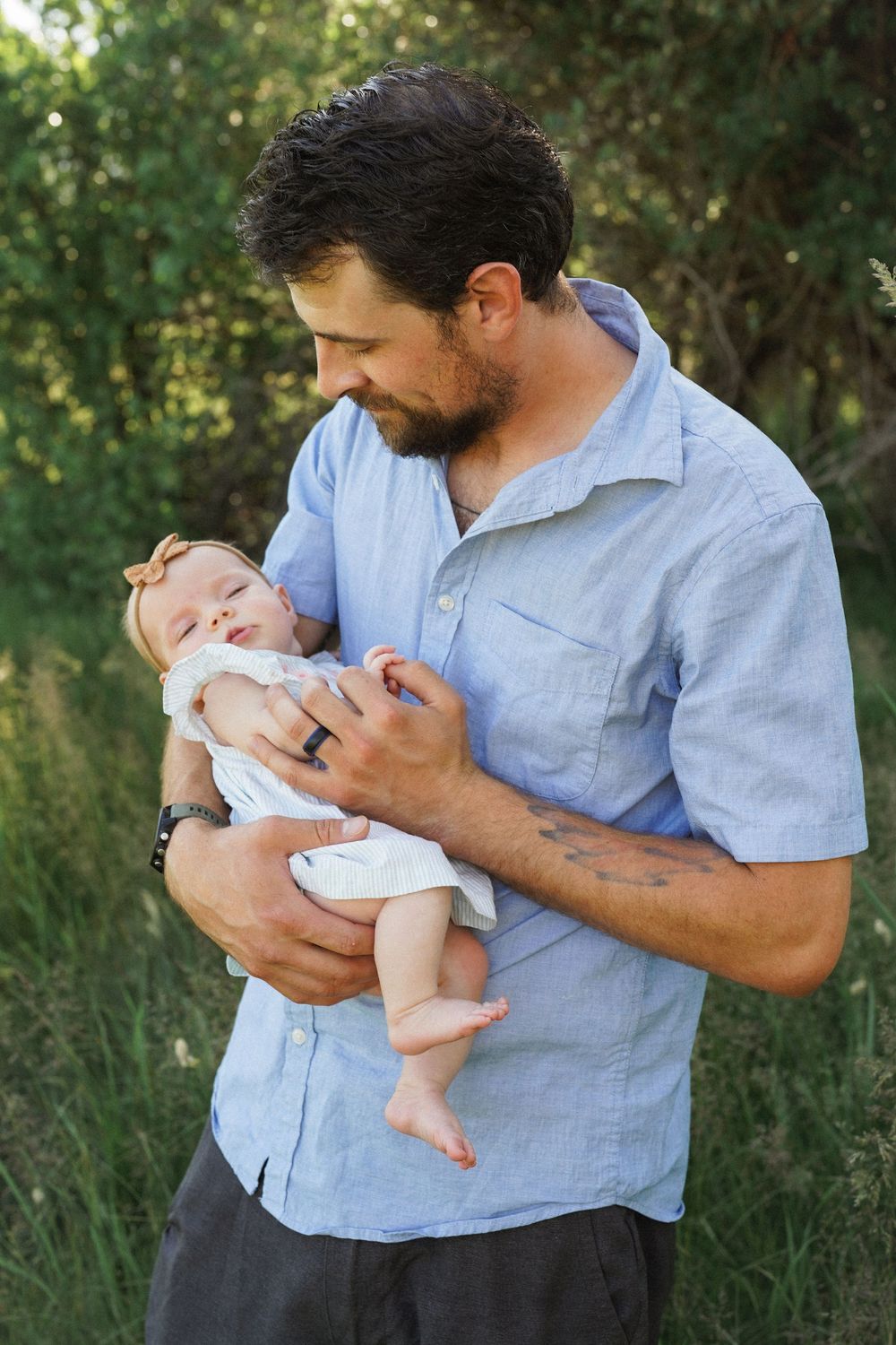 A person in a blue shirt holds and feeds a small baby outdoors in a natural setting with greenery in the background.