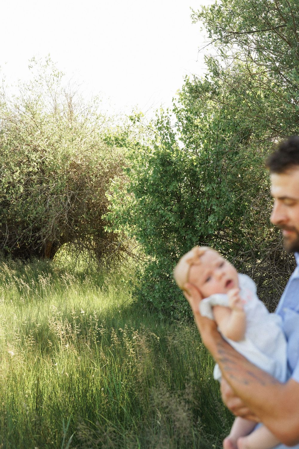 A tender moment captured outdoors with a baby being held in sunlit greenery and tall grass.