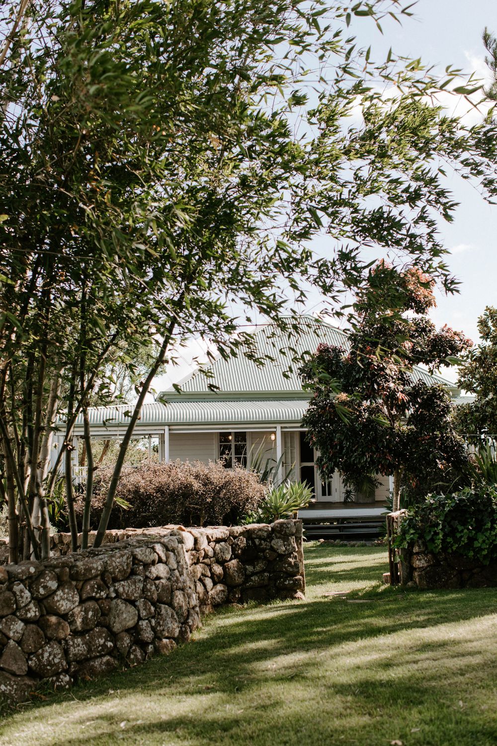 Classic white farmhouse with stone wall and manicured gardens viewed through overhanging trees.