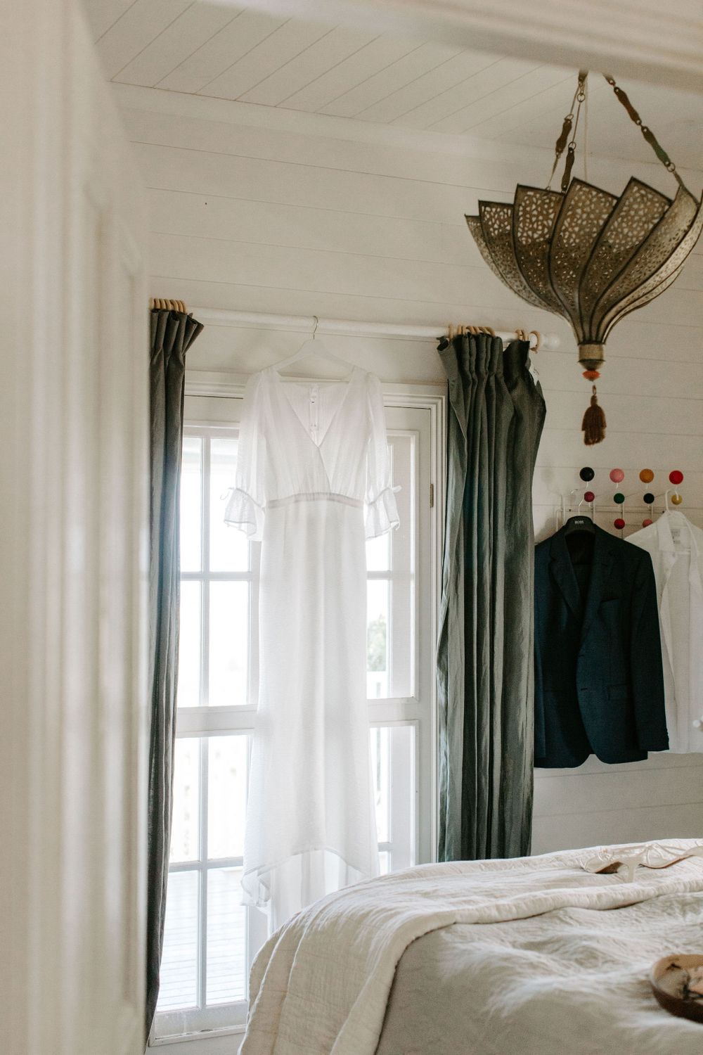 White wedding dress and navy suit hanging by windows with green curtains and decorative chandelier.