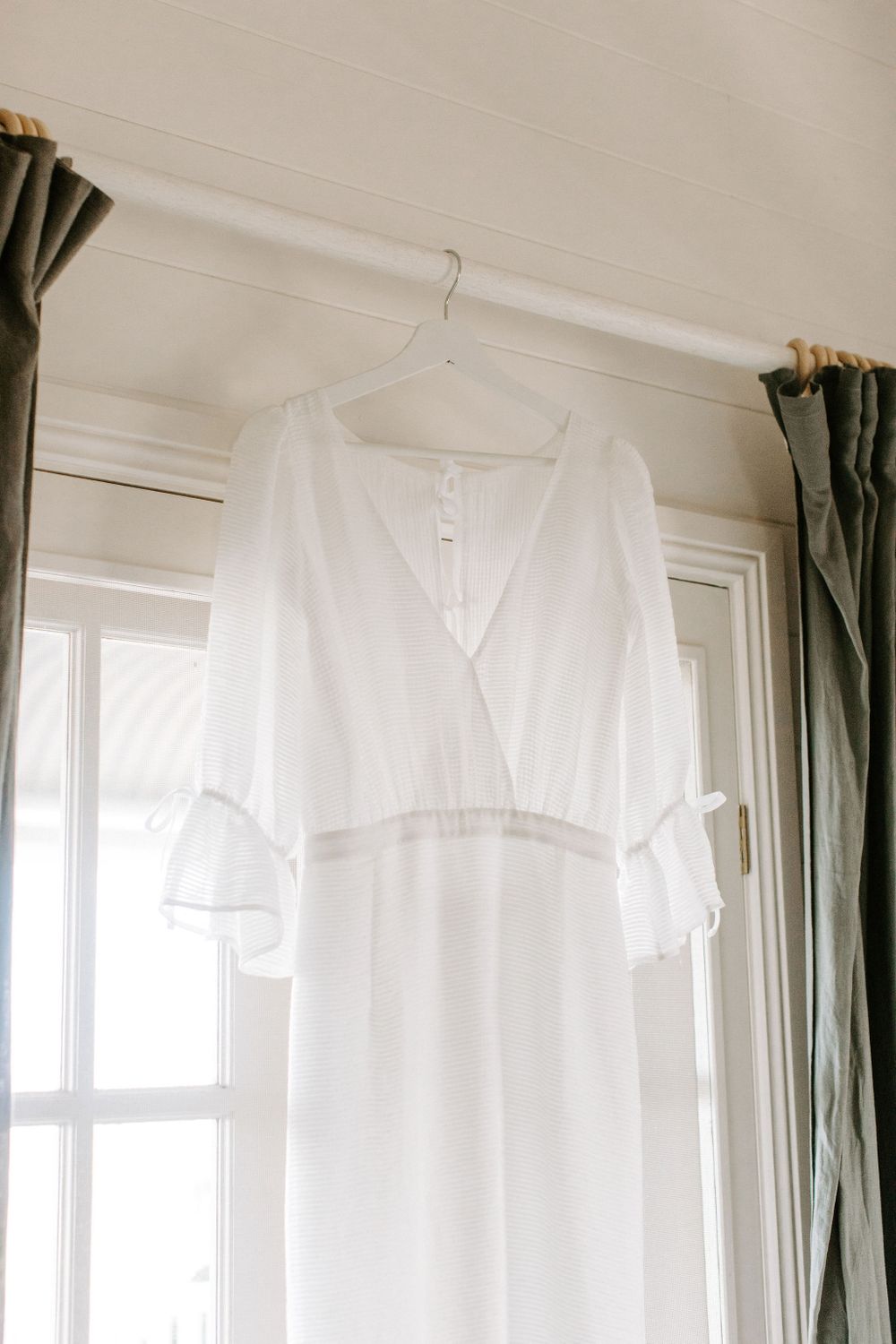 Elegant white wedding dress with ruffled sleeves hanging against a bright window.