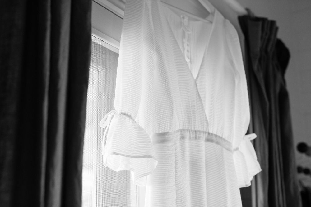 Black and white photo of a delicate white wedding dress hanging by a window.