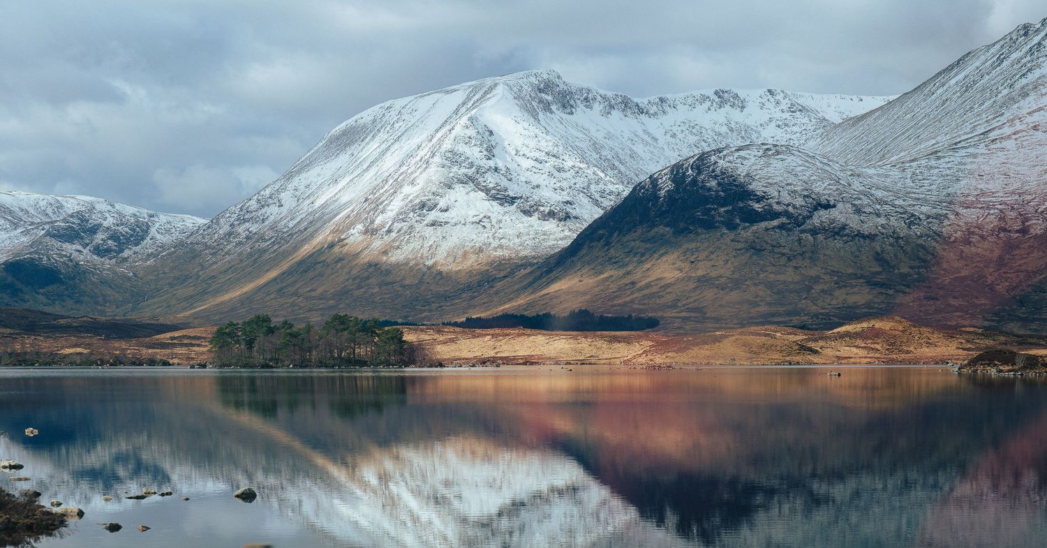 Snow-capped mountains reflect perfectly in a calm lake under moody winter skies.