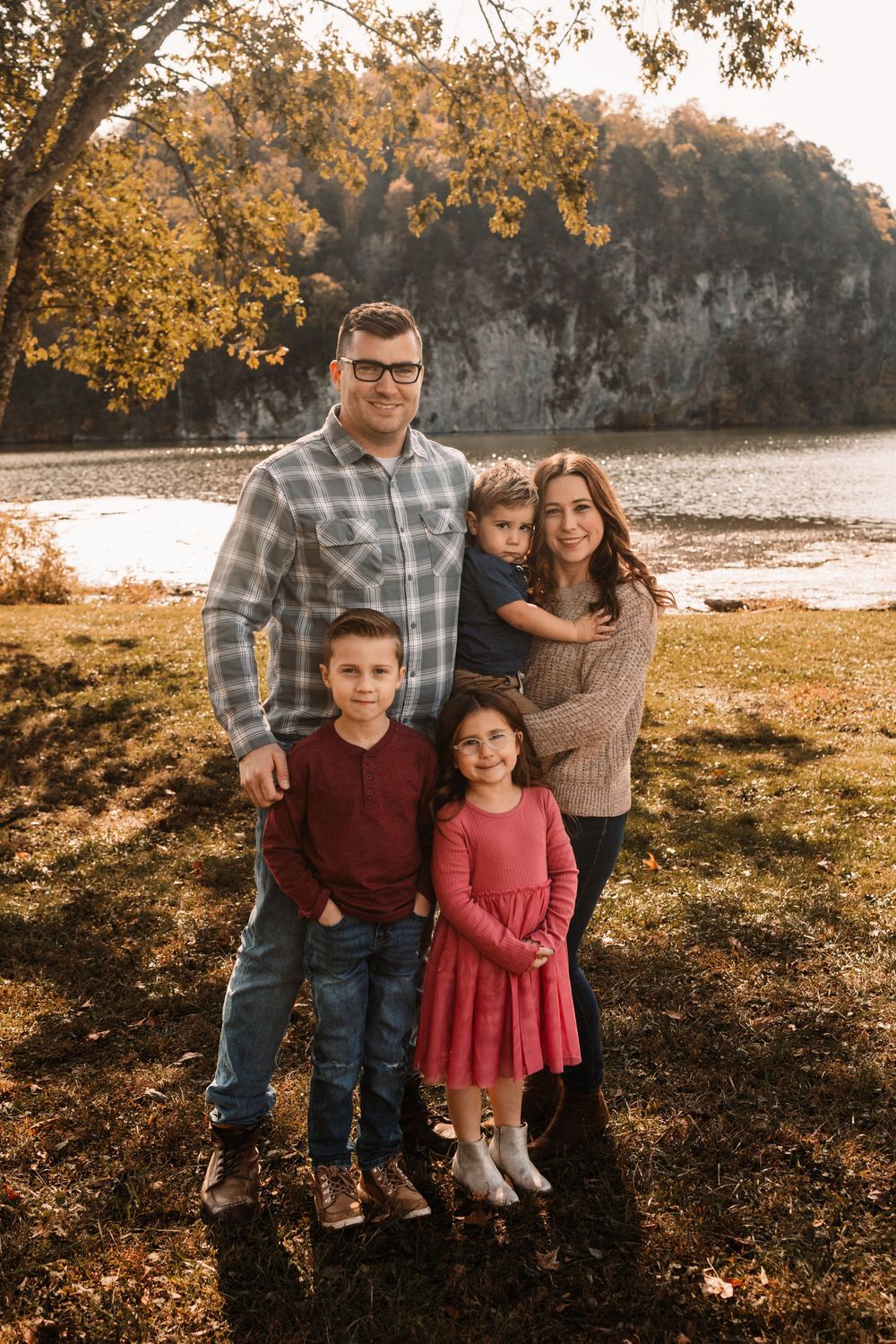 Family of five poses by a rocky riverside in coordinated fall outfits at sunset.