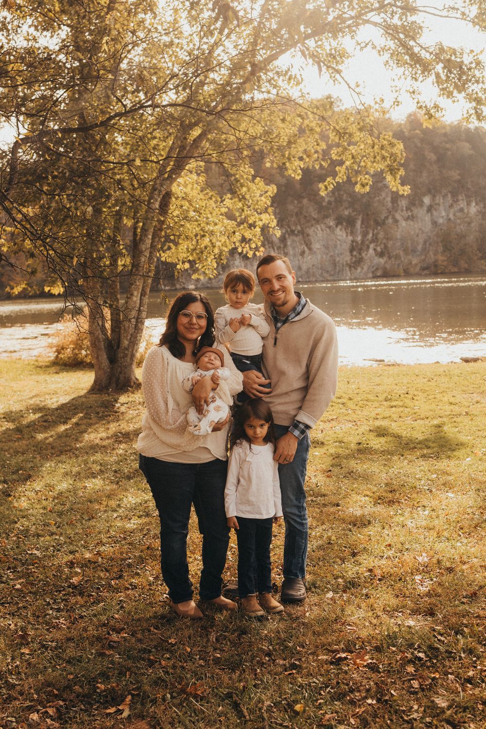 Happy family of five poses under golden autumn tree near a peaceful river during fall season.