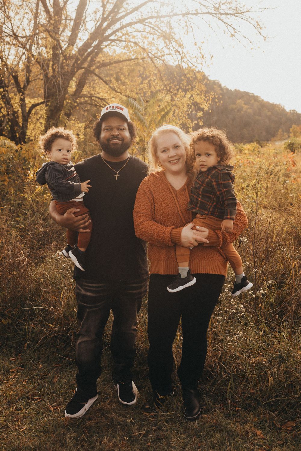 Family of four poses in autumn field, parents holding two toddlers in warm fall colors.