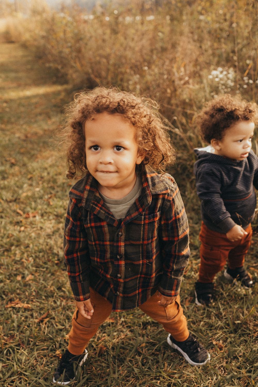 Two young children with curly hair stand in a sunlit autumn meadow wearing plaid and rust outfits.