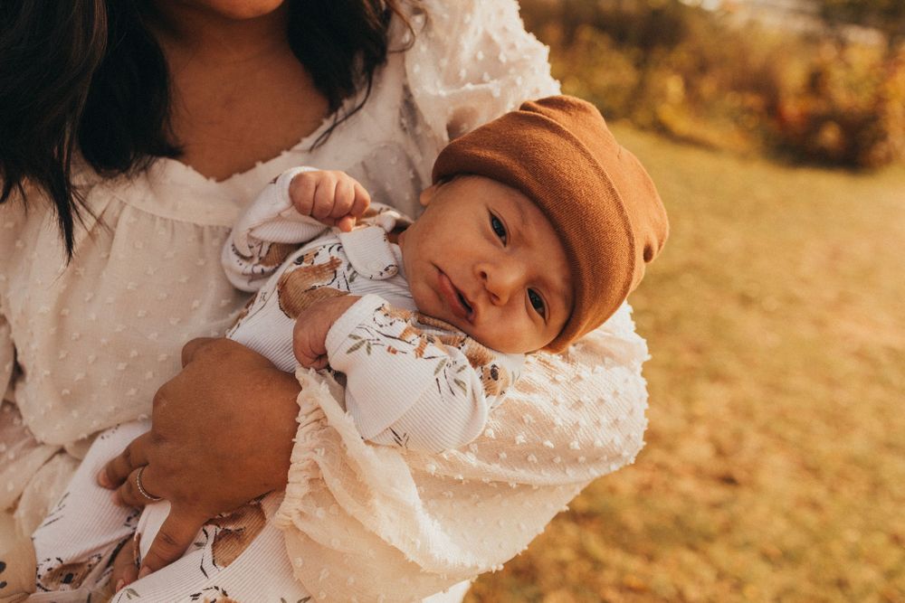 Mother tenderly holds newborn baby dressed in white with a brown hat in outdoor autumn setting.