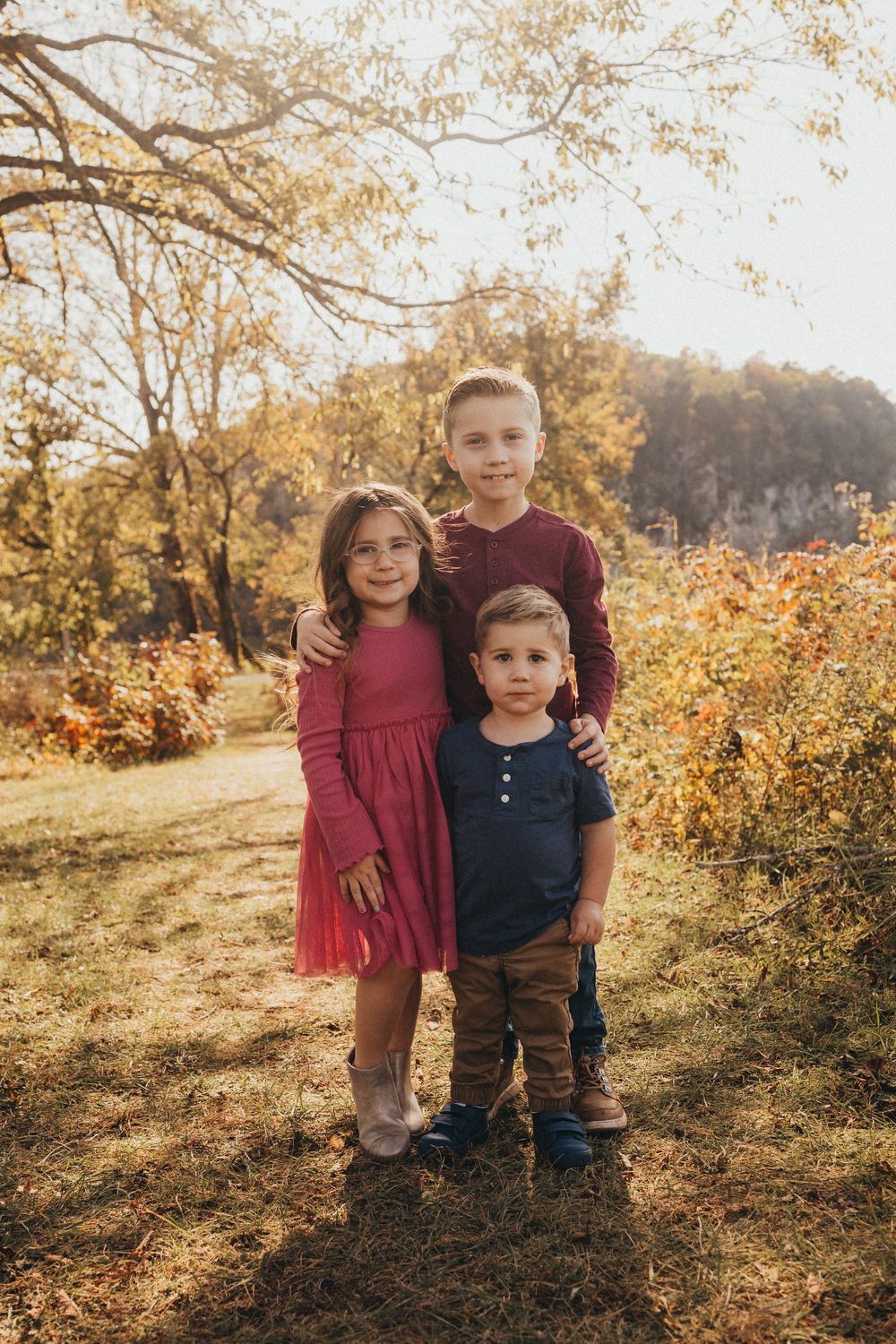 Three siblings embrace outdoors in golden autumn light, wearing coordinated fall outfits.