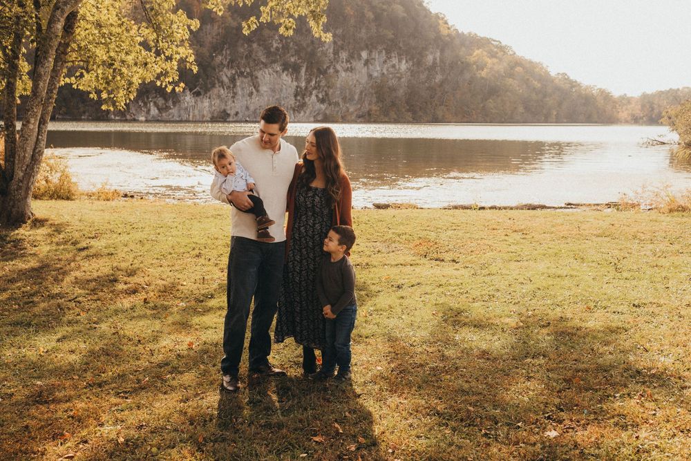 Family of four stands together near a scenic river surrounded by autumn foliage.
