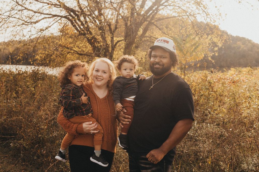 Smiling family of four poses together in golden autumn field near a lake at sunset.