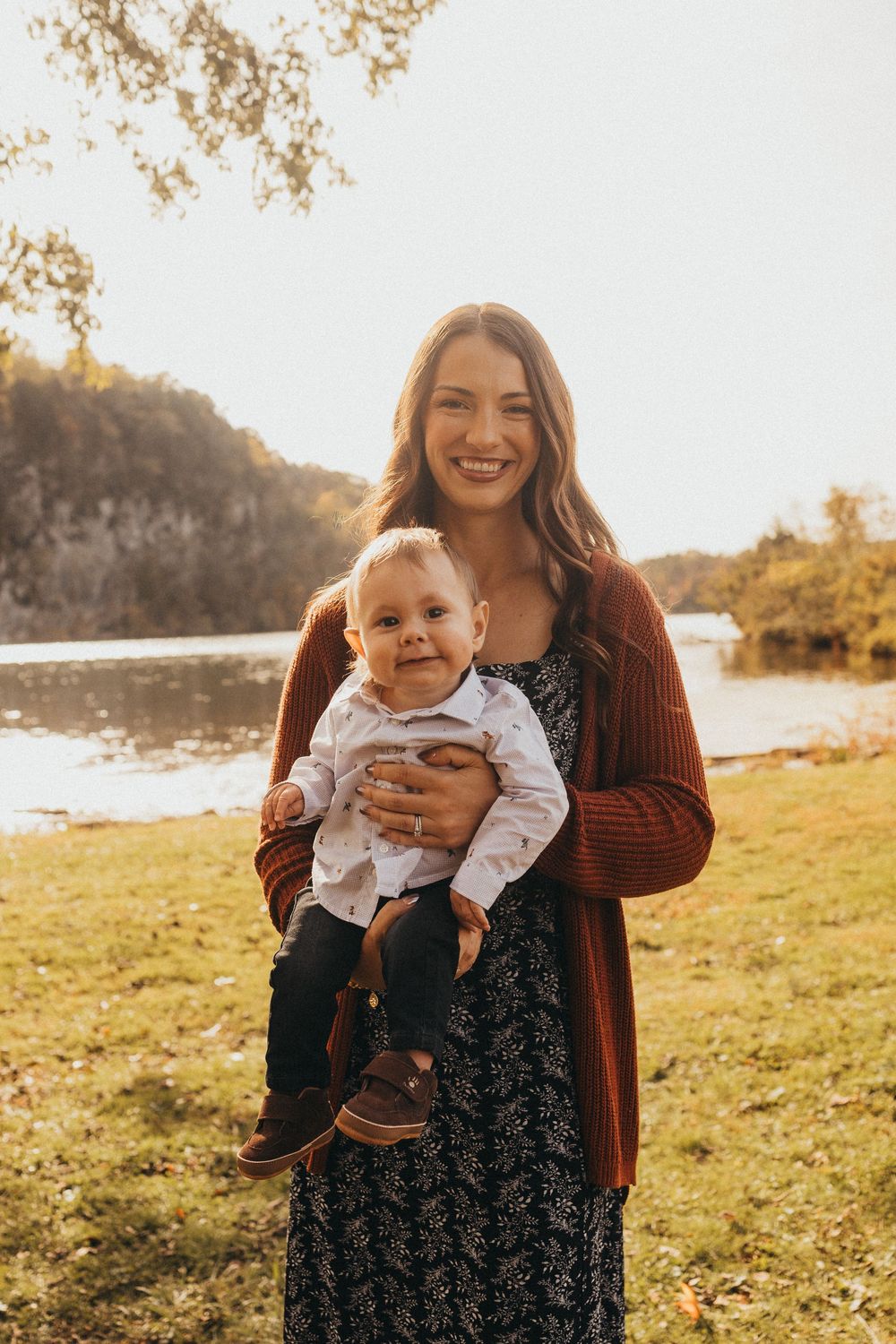Smiling mother holds happy baby outdoors by a river in golden autumn sunlight.