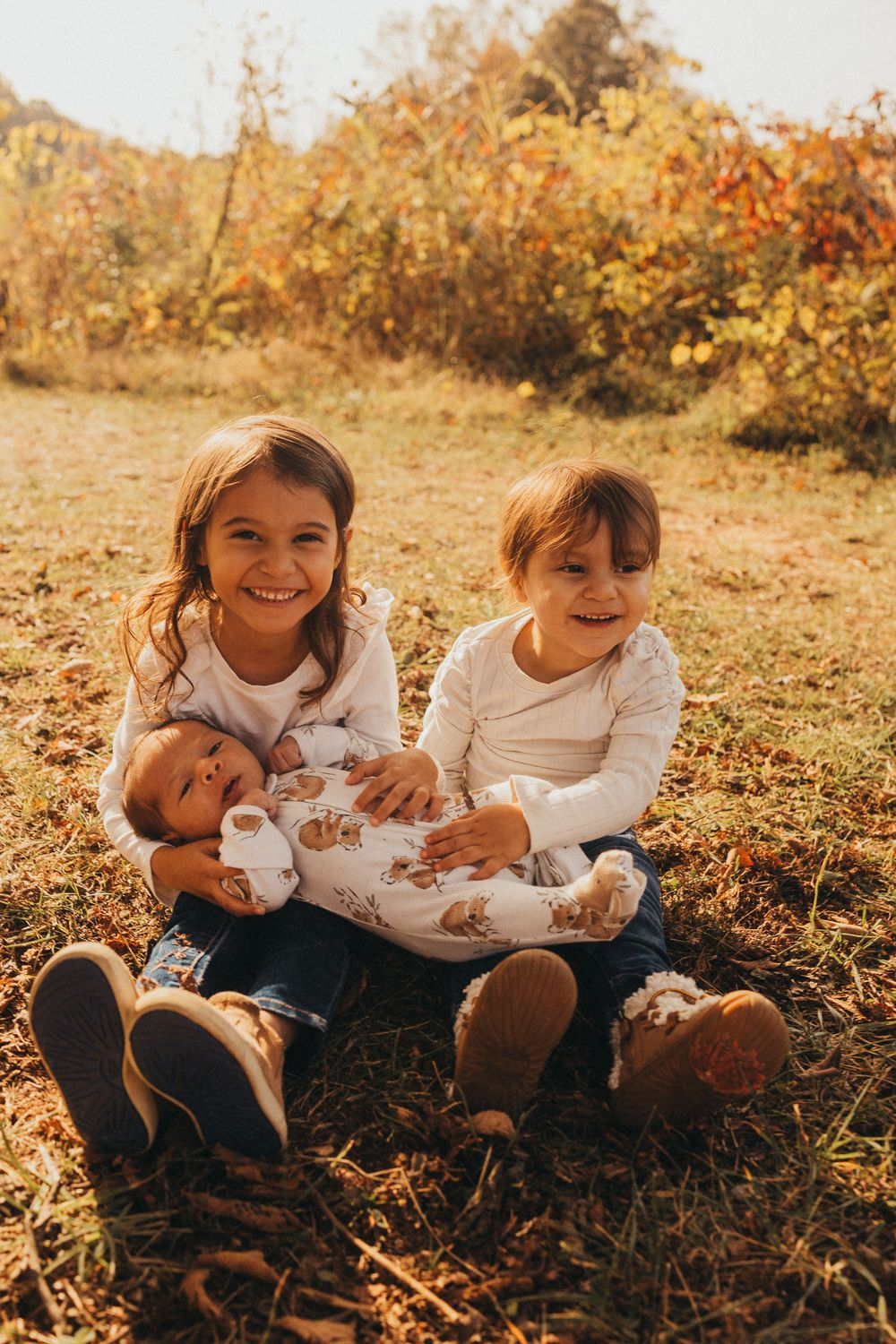 Two smiling siblings sit on autumn grass holding their newborn baby sibling in warm sunlight.