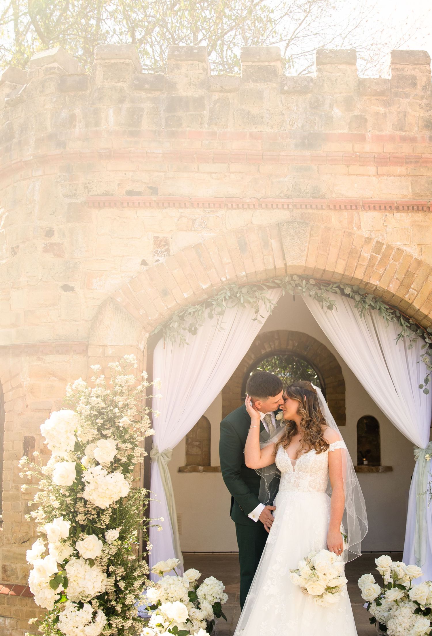 A romantic wedding moment captured under elegant white-draped stone archways adorned with white floral arrangements.