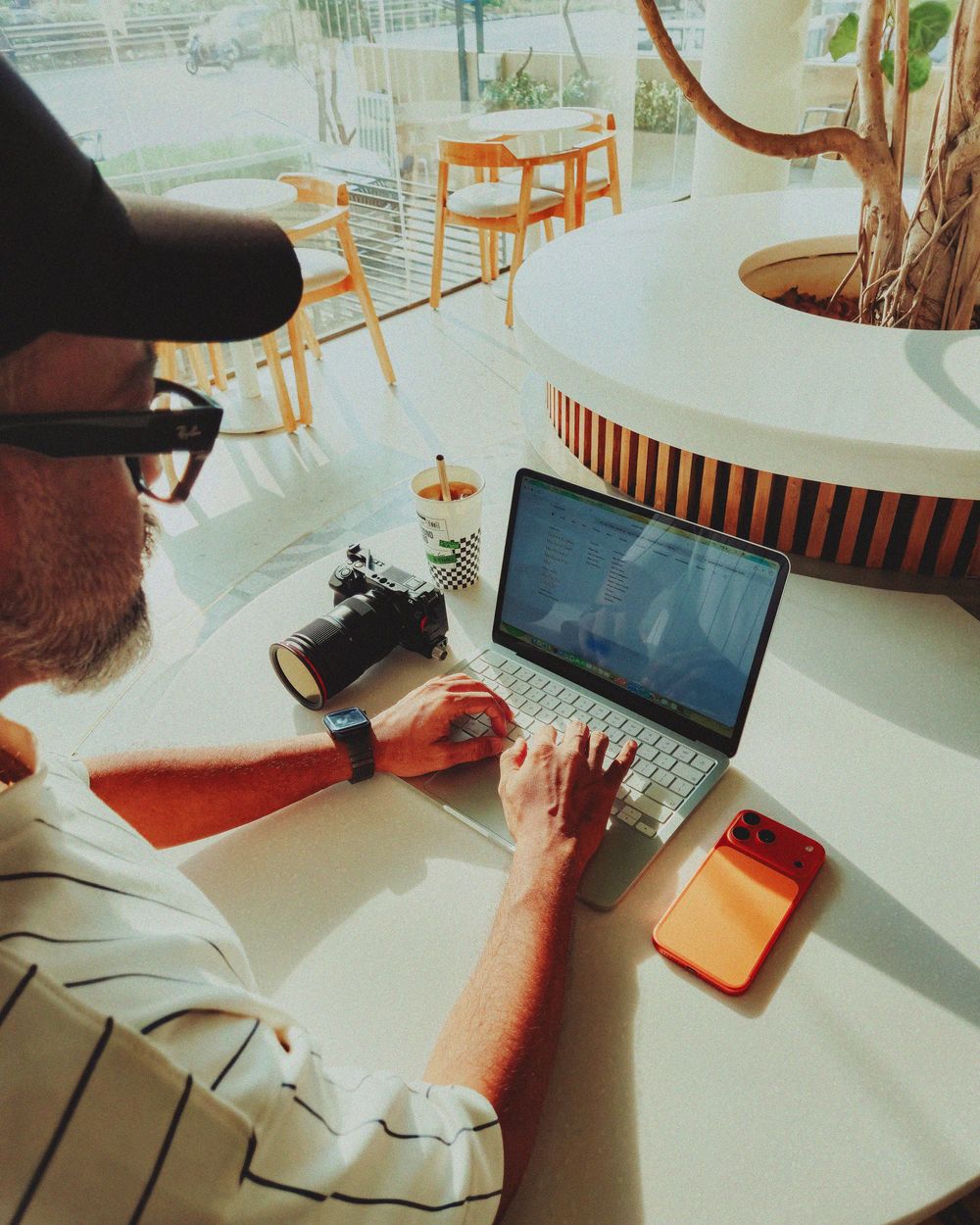 Man working on laptop at white cafe table with camera, smartwatch, and orange phone nearby.