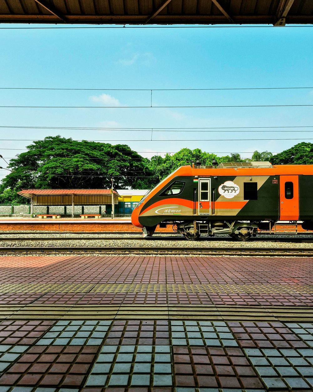 Orange modern train at a tropical railway station platform with lush green trees and blue sky in the background.