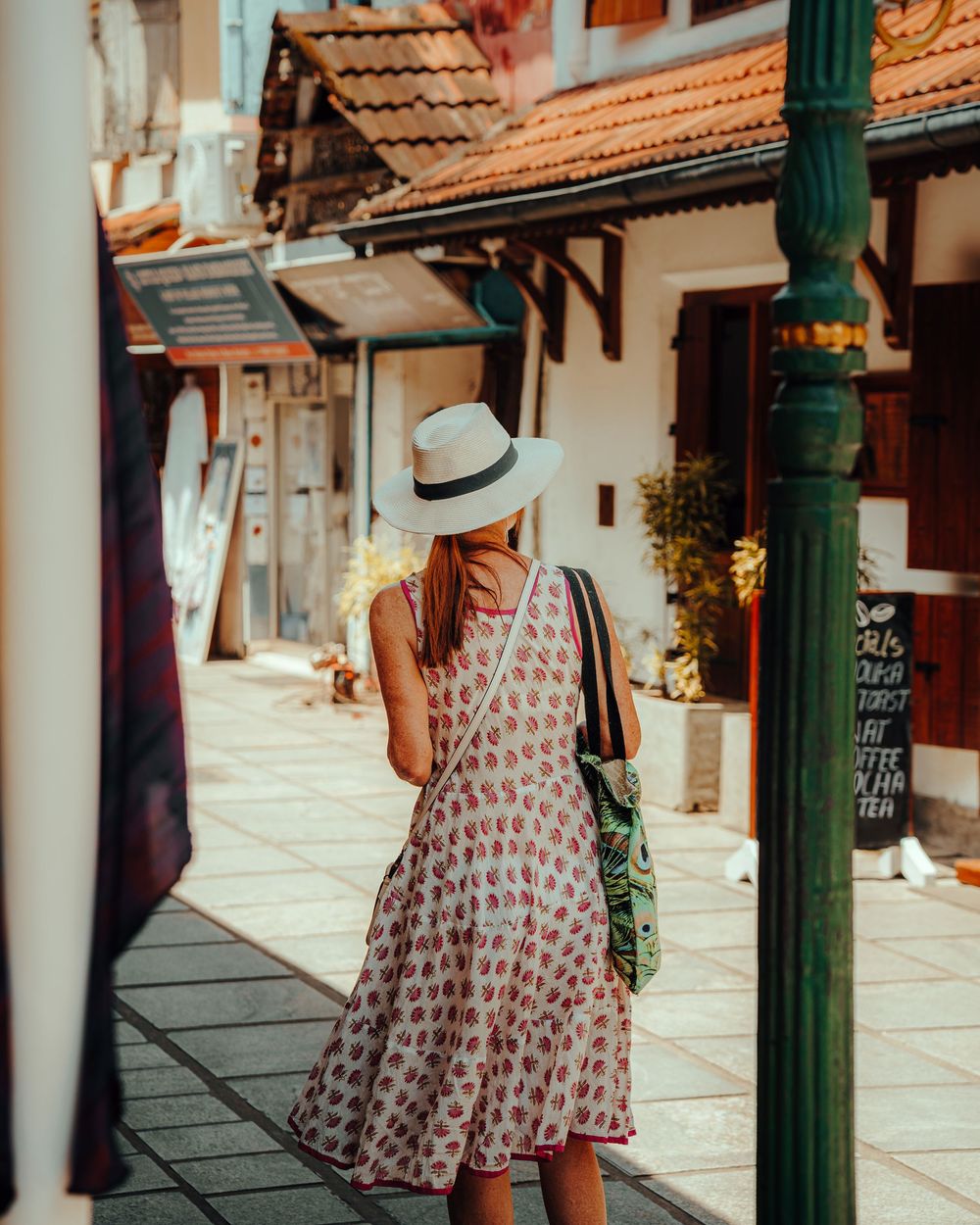Woman in a white sun hat and pink floral sundress walks down a sunlit cobblestone street lined with charming terracotta-roofed buildings.