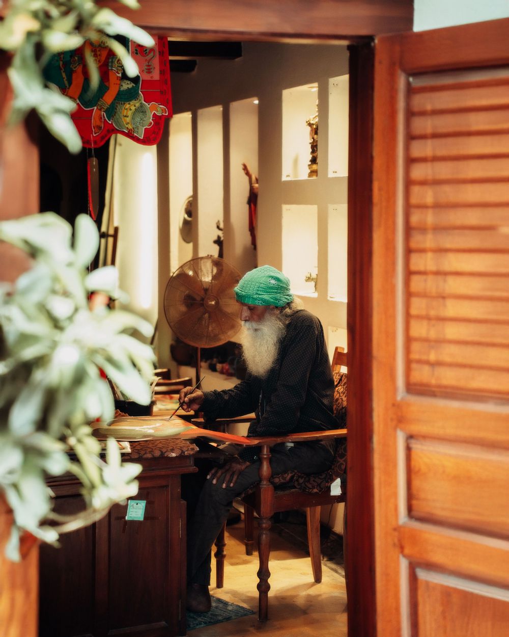 Through a doorway, a person in green headwear sits at a wooden desk in a warm-lit room with wooden shutters.
